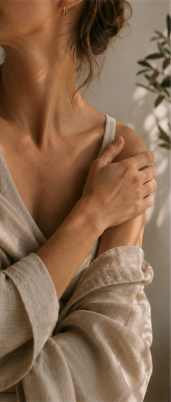Mujer sosteniendo suavemente su hombro en un gesto de autocuidado y pausa consciente, iluminada por luz natural cálida.